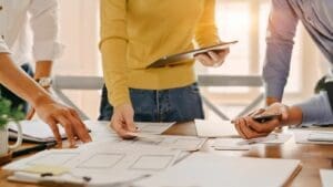 Hands and torso of three designers holding mobile devices while leaning over table with website planning documents