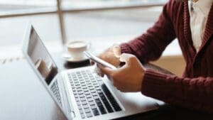 Close up on man's hands holding cell phone while in front of open laptop at a cafe table with coffee and a window in the background.