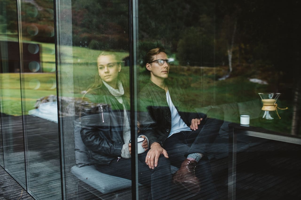 Two people seated indoors behind a large glass window, looking outward at a grassy landscape; reflections of trees and outdoor scenery appear on the glass, with a small table, coffee cup, and carafe visible beside them.